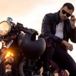 Handsome rider guy with beard and mustache in black cafe racer jacket take off sunglasses on classic style cafe racer motorcycle at sunset.