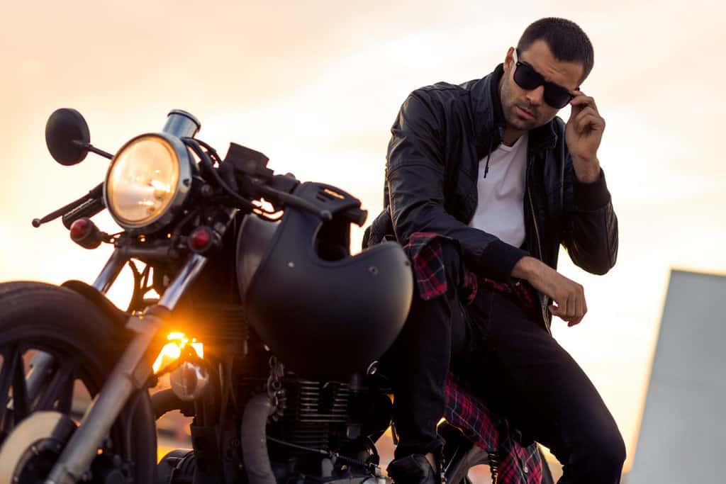 Handsome rider guy with beard and mustache in black cafe racer jacket take off sunglasses on classic style cafe racer motorcycle at sunset.