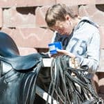 Teenage girl equestrian cleans black leather horse saddle and equipment at farm.